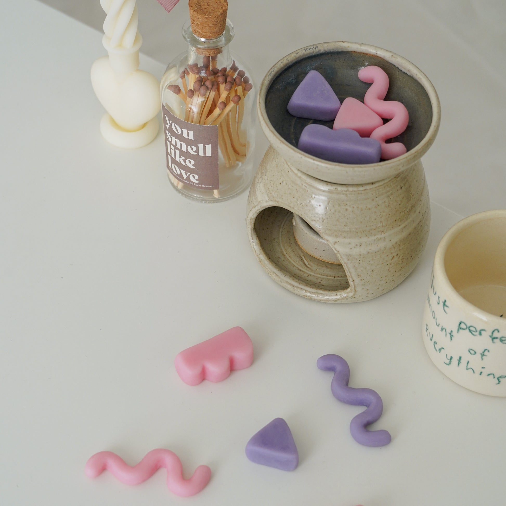 pastel geometric shape unique wax melts in a wax warmer, a match bottle, a heart twist candle, and a mug on white table