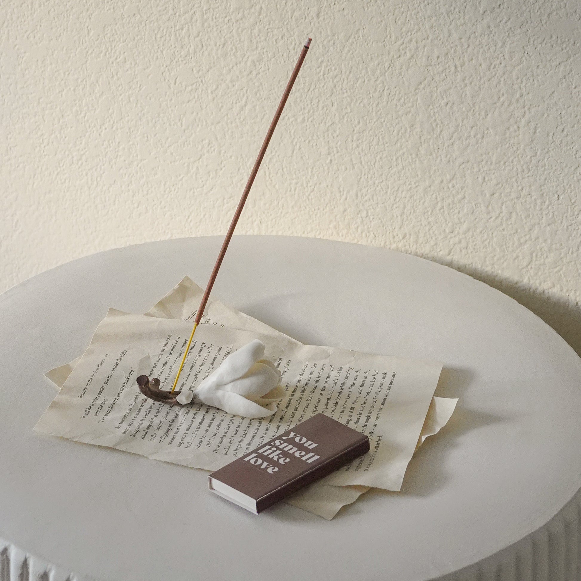 magnolia flower incense holder with an incense stick on book pages and a brown match box inscribed with you smell like love on white end table