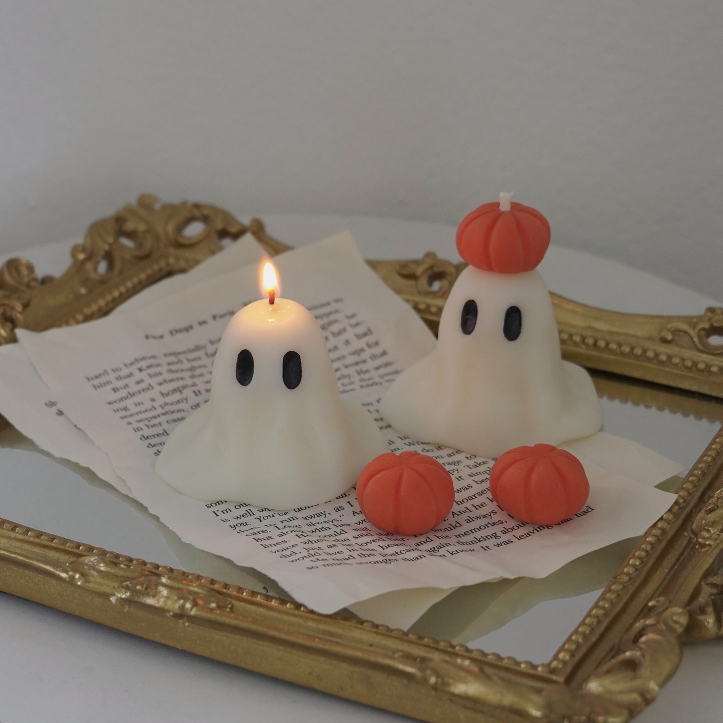 a lit ghost candle, pumpkin ghost candle, and pumpkin wax melts on book pages and rectangular gold french mirror tray