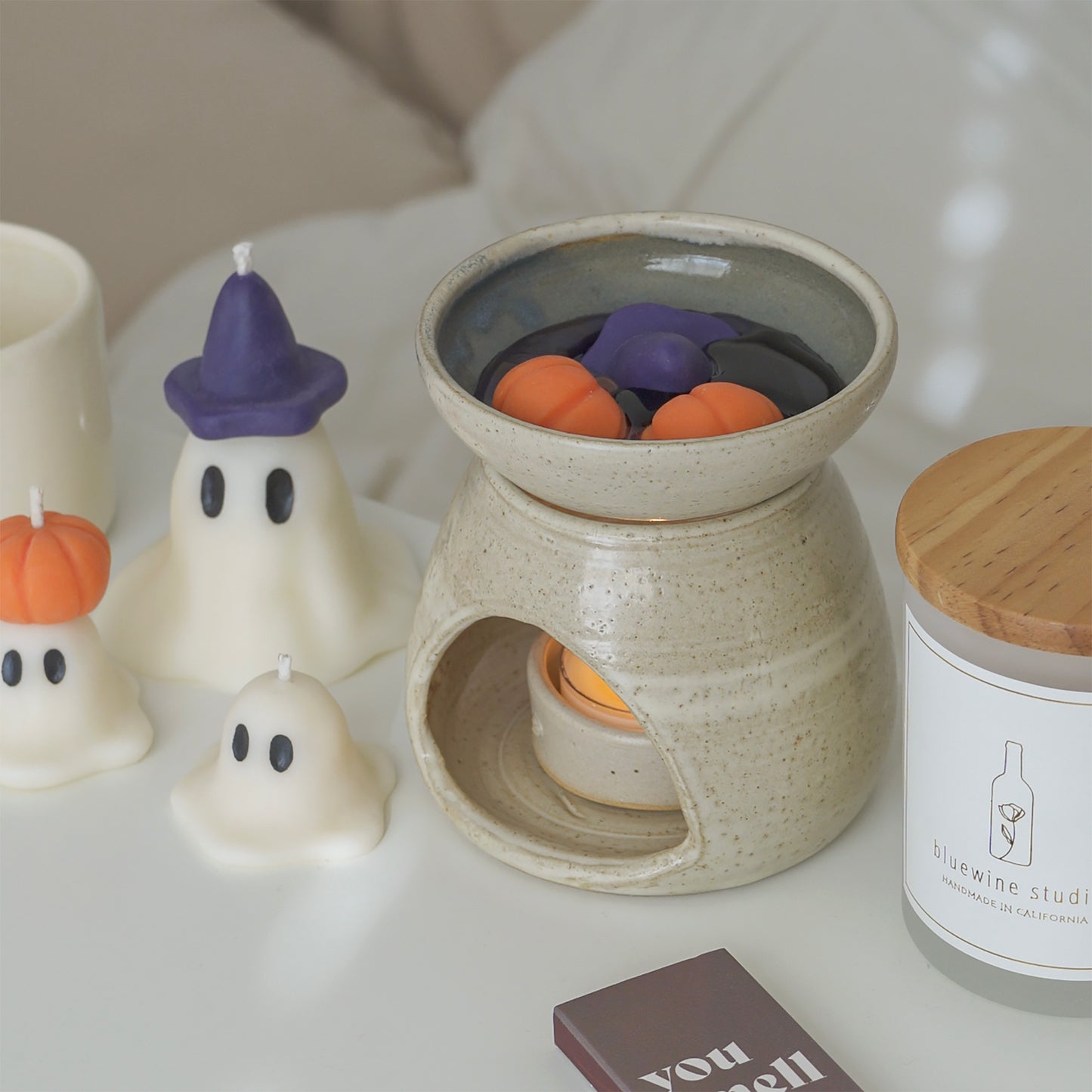 purple wizard hat and pumpkin wax melts melting in a wax warmer, a container soy candle with wooden lid, brown matchbox, and wizard ghost candle on a white table