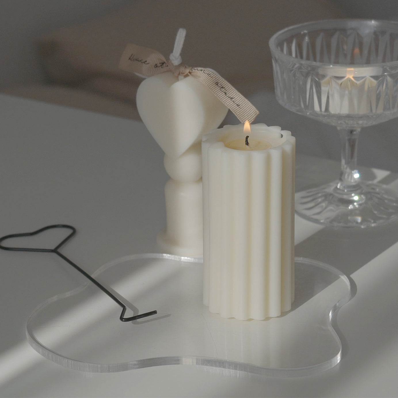 Heart shape candle, a burning ribbed pillar candle on a clear irregular shape acrylic coaster with a black metal heart shaped wick dipper, and a lit tealight candle in a glass coupe are placed on white drawer. The arrangement exudes minimal aesthetic dreamy vibes.