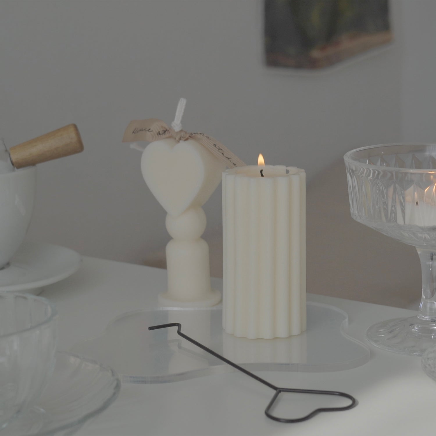 Heart shape candle, a burning ribbed pillar candle on a clear irregular shape acrylic coaster with a black metal heart shaped wick dipper, and a lit tealight candle in a glass coupe are placed on white drawer. The arrangement exudes minimal aesthetic dreamy vibes