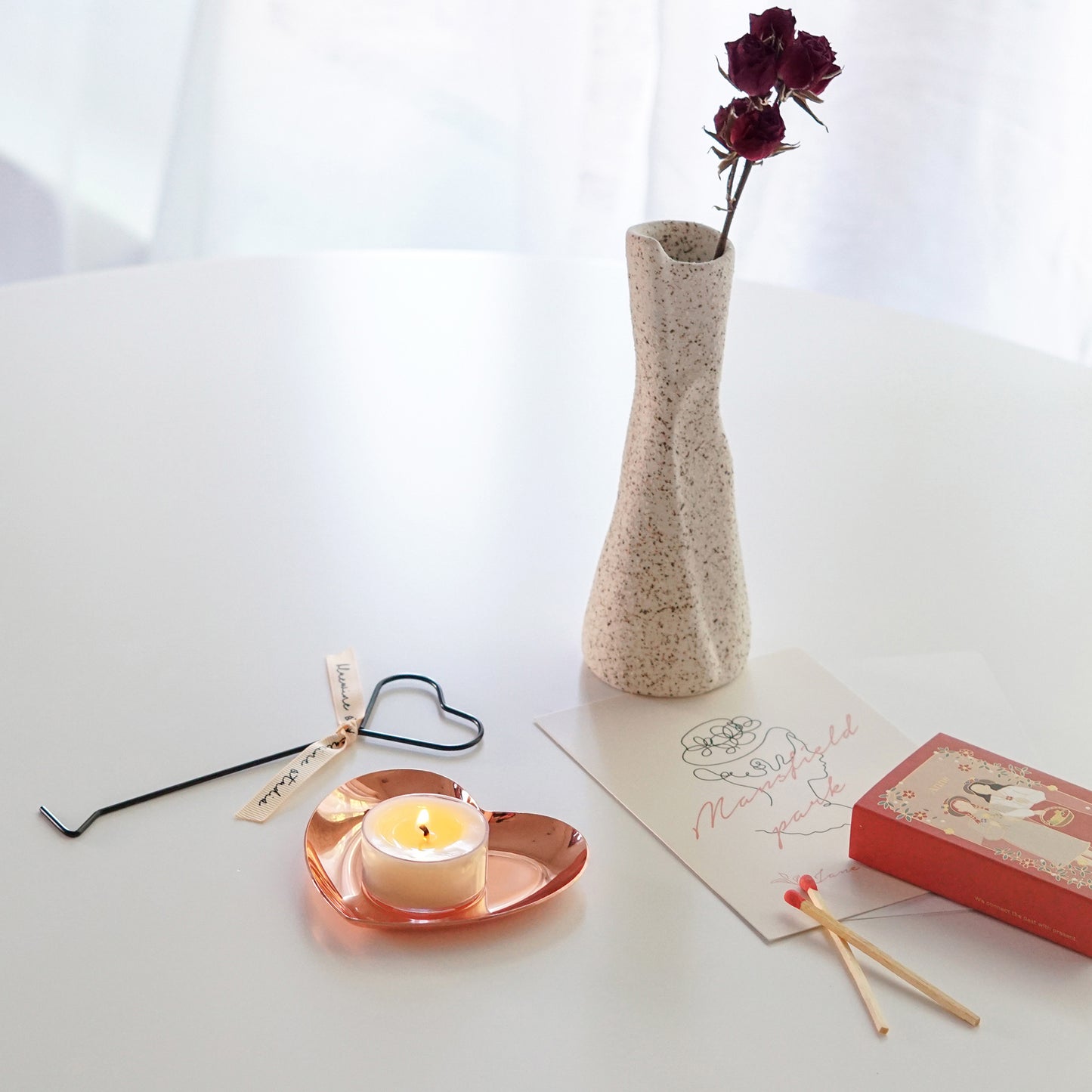 a lit tealight candle on rose gold heart tray, heart wick dipper, red matches, one line drawing minimal postcard, and burgundy dried roses in ceramic unique vase on white round table