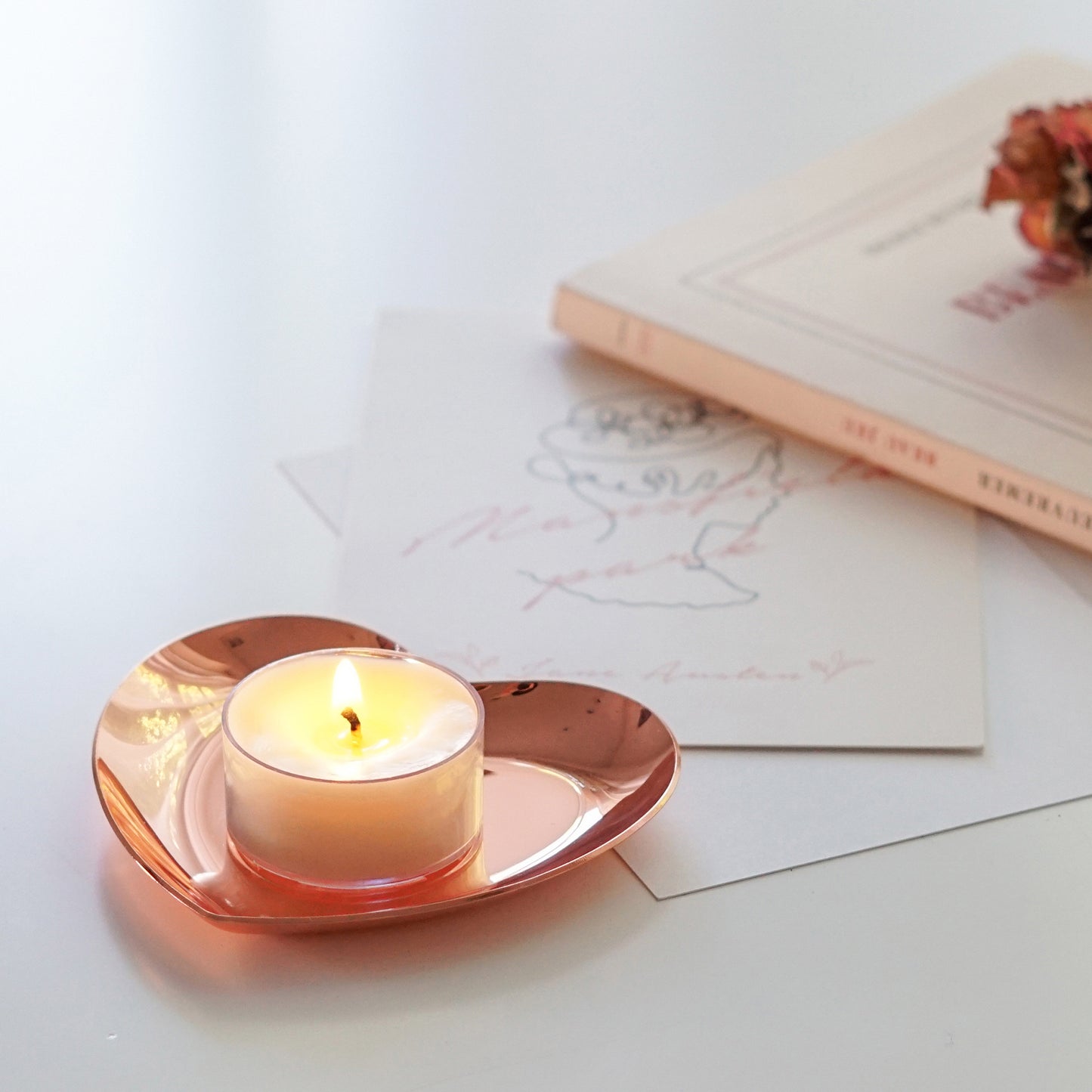 a lit tealight candle on rose gold heart tray, postcards, french book, and pink dried roses