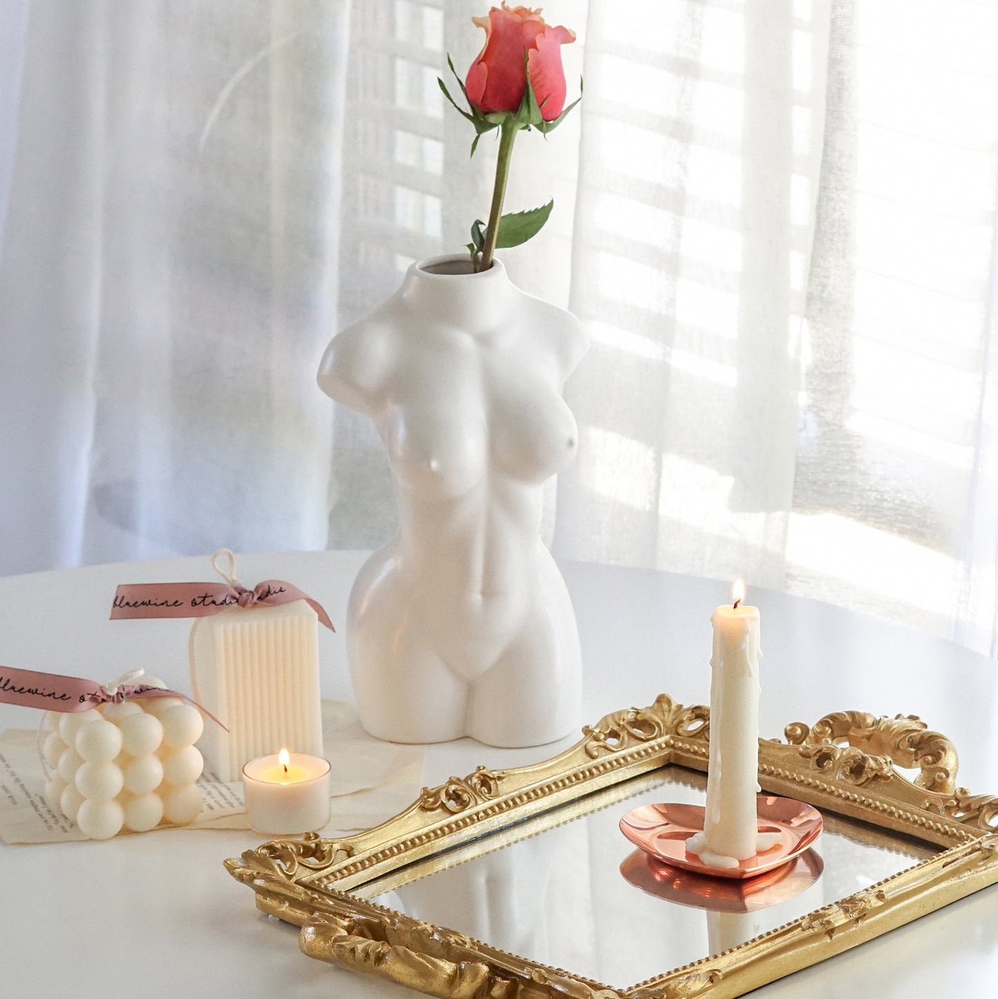 a lit taper candle on rose gold heart shape tray placed on rectangular gold french mirror tray, cube bubble soy pillar candle and ribbed square pillar candle with pink bluewine studio ribbons, a lit tealight candle, book pages, and a pink rose in a white ceramic feminine body shape vase