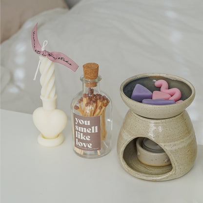 a heart twist taper soy candle, a match bottle with a brown sticker inscribed with you smell like love, and a pastel unique geometric shape wax melts in a wax warmer on the white table