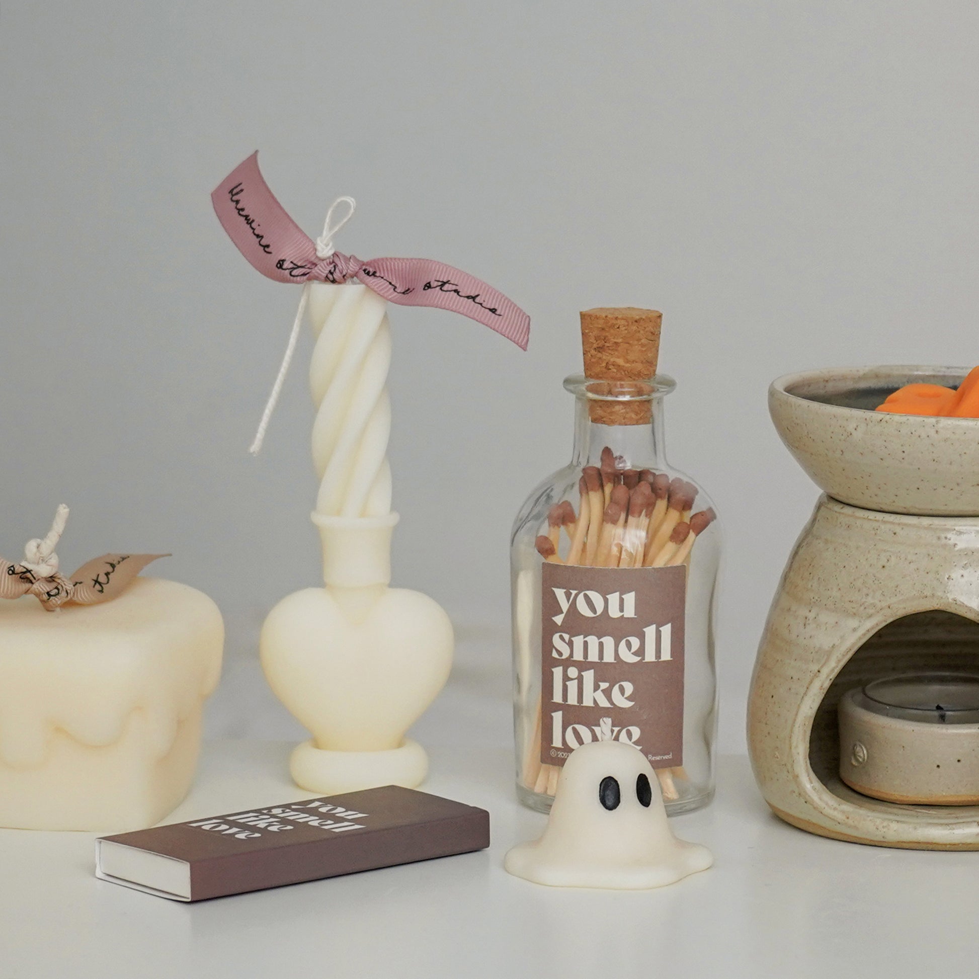a heart twist taper candle, a mini ghost candle, a brown matchbox, a match bottle, and cheese wax melts in a wax warmer on the white table