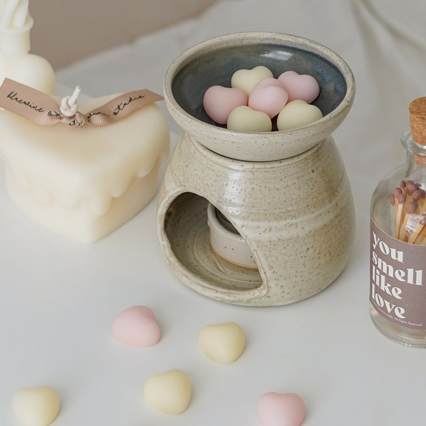 a melting heart candle, white and pink wax melts in a wax warmer, and a match bottle with a brown sticker inscribed with you smell like love on the white table