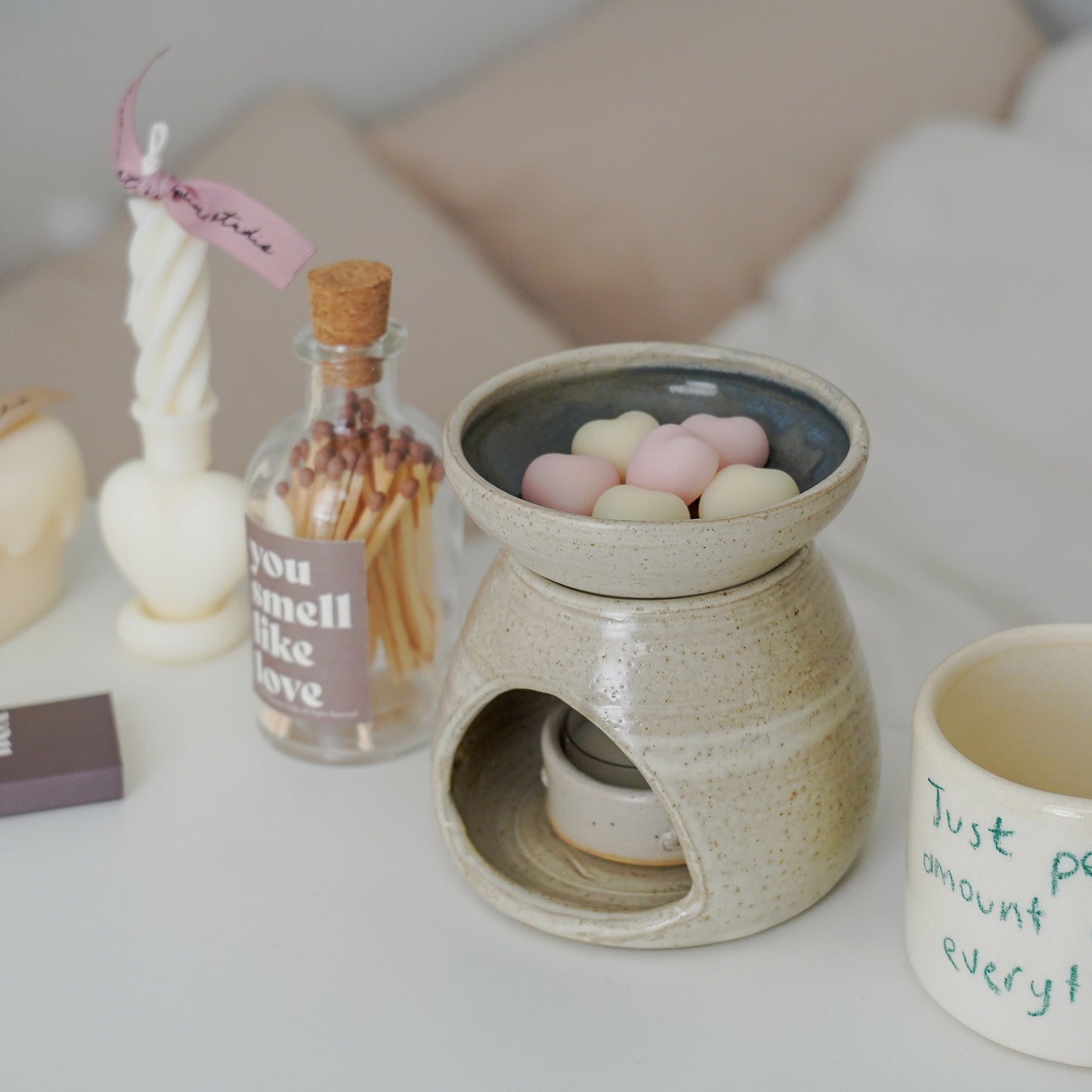 a match bottle with brown sticker inscribed with you smell like love, pink and white heart wax melts in a wax warmer, a ceramic mug, and a heart twist taper candle on the white table