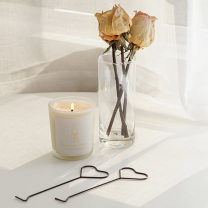 two black heart shape candle wick dippers, a lit bluewine studio frosted glass container soy wax candle, and two yellow dried roses in a clear vase