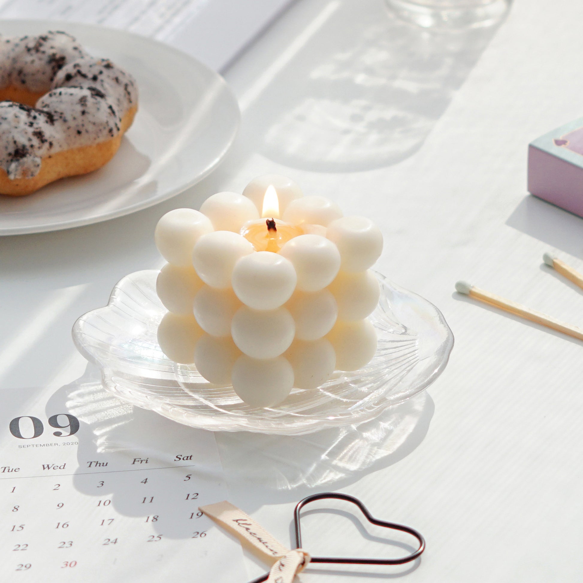 a lit white square cube bubble soy pillar candle on a holographic shell tray, september calendar postcard, heart wick dipper, mint color matches, and oreo cookie mochi donut on a white ceramic plate