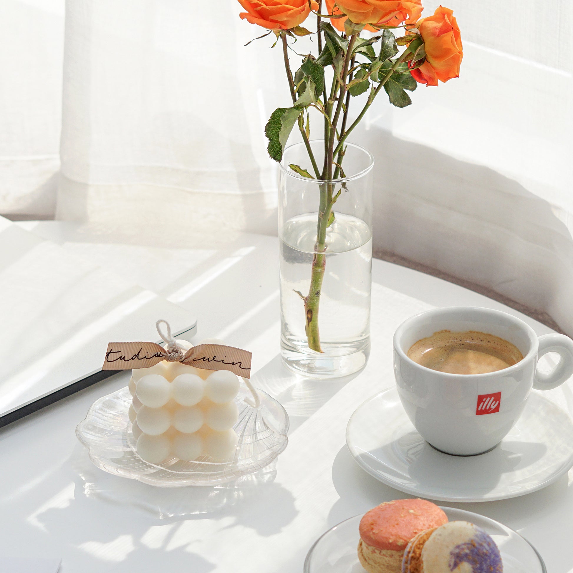 a white square cube bubble soy pillar candle with beige bluewine studio ribbon on a holographic shell tray, hot coffee in illy mug set, orange roses in clear transparent cylider vase, silver macbook, and two macarons on a clear dish are on white round table