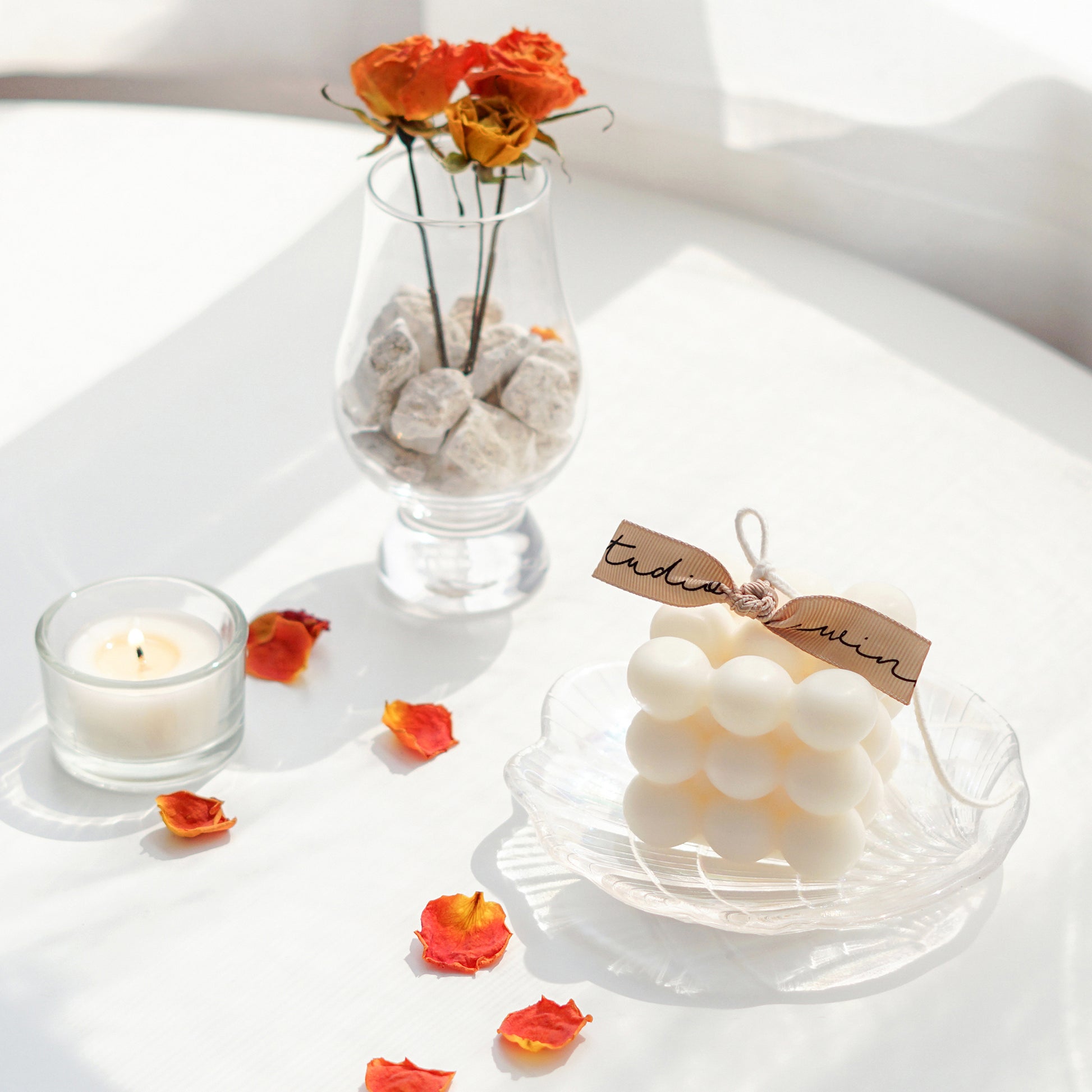 a white cube square bubble soy pillar candle on a holographic shell tray, a lit tealight candle in a tealight glass holder, orange dried roses in a brandy glass with white rocks, and rose petals on white round table