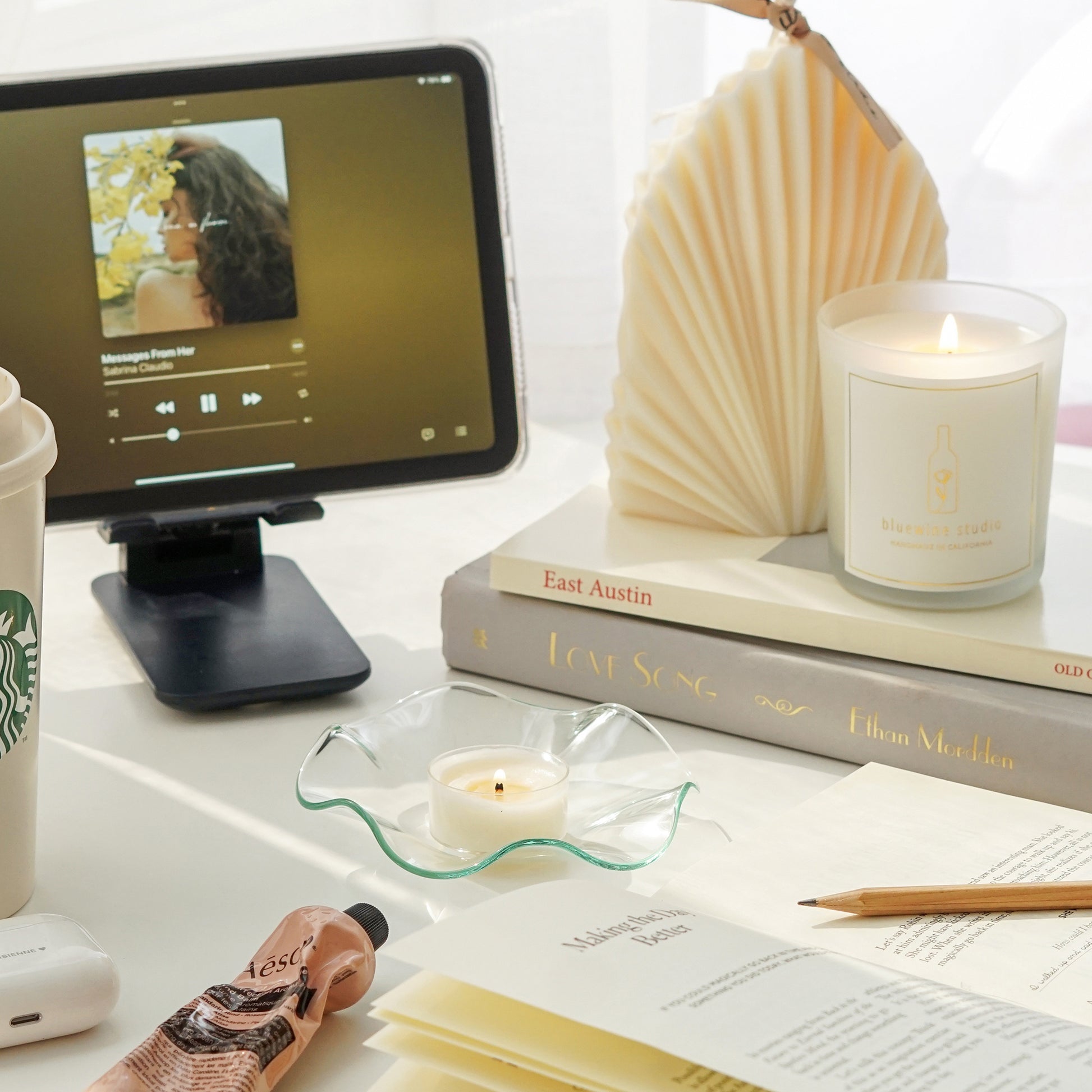 a leaf shape sculptural white natural soy pillar candle and a lit 5oz frosted glass container candle on book stacks, ipad mini playing music messages from her by sabrina claudio, the five minute journal with pages open, a lit tealight candle on a clear wavy ruffle dish, aesop hand lotion, airpods, and starbucks tumbler