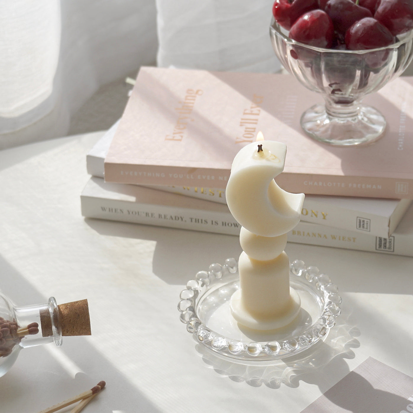 a lit crescent moon shape candles and cherries in a clear ice cream bowl on stacked books are placed on white table. The arrangement exudes minimal aesthetic dreamy vibes.