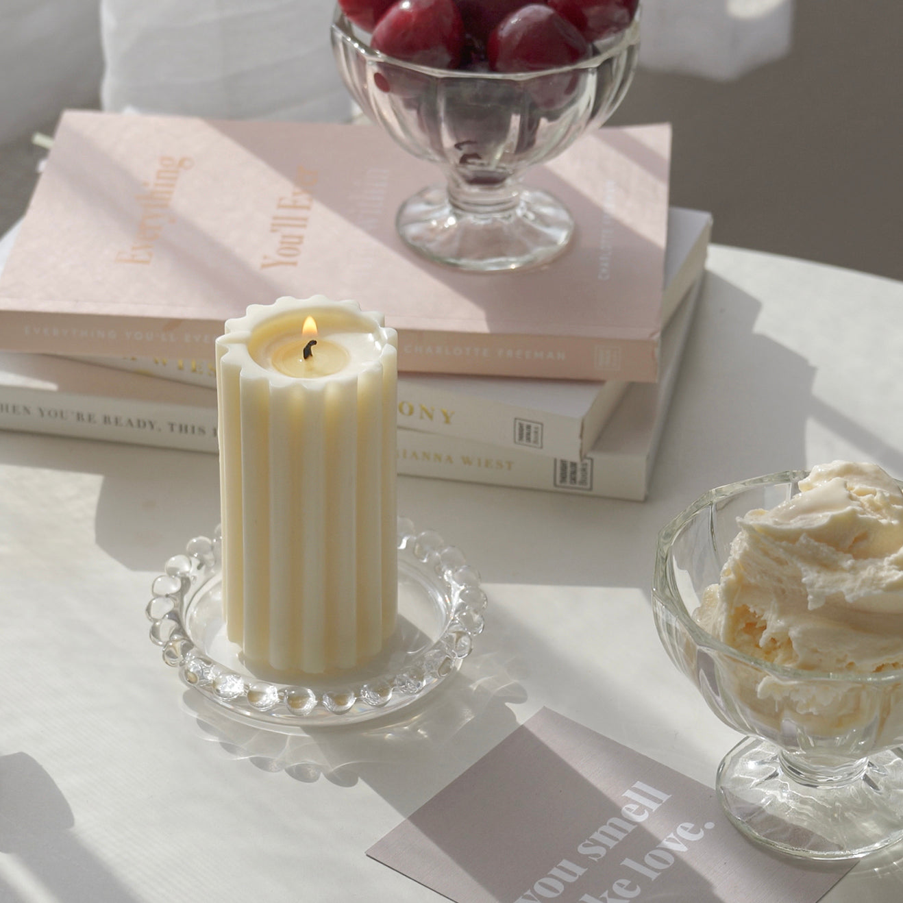a lit ribbed pillar soy candle on a clear mini beaded tray, ice cream in a bowl, postcard, books, and cherries in an ice cream bowl on white round table