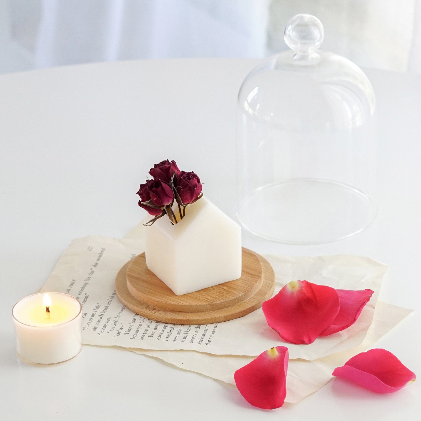 mini house white scented wax with burgundy dried roses on round wood coaster, rose petals, and a lit tealight candle placed on book pages