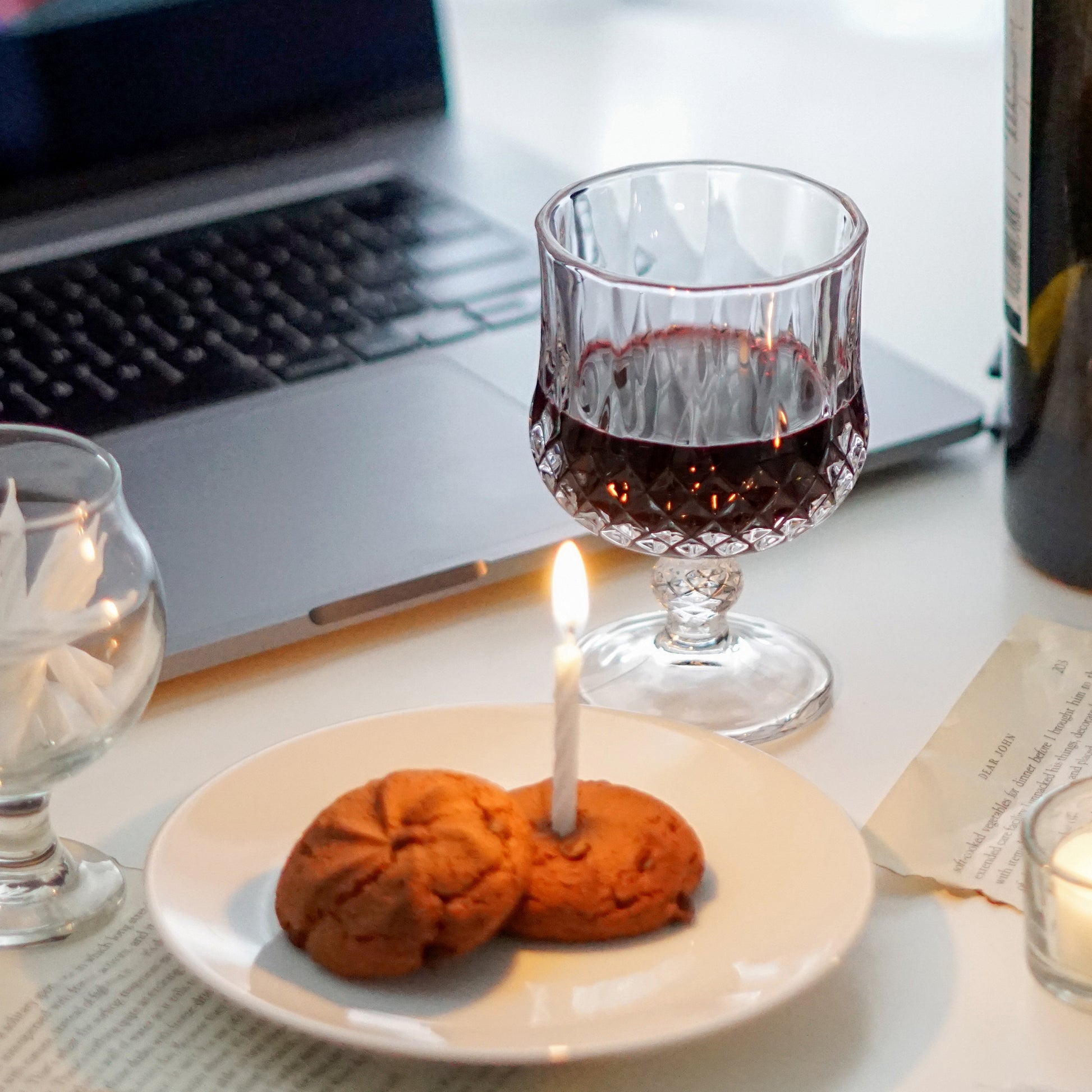 red wine in a mini wine glass, cookies on ceramic white dish, mini cake candles in a glass, a lit tealight candle in a glass candle holder, a bottle of wine, book pages, and macbook