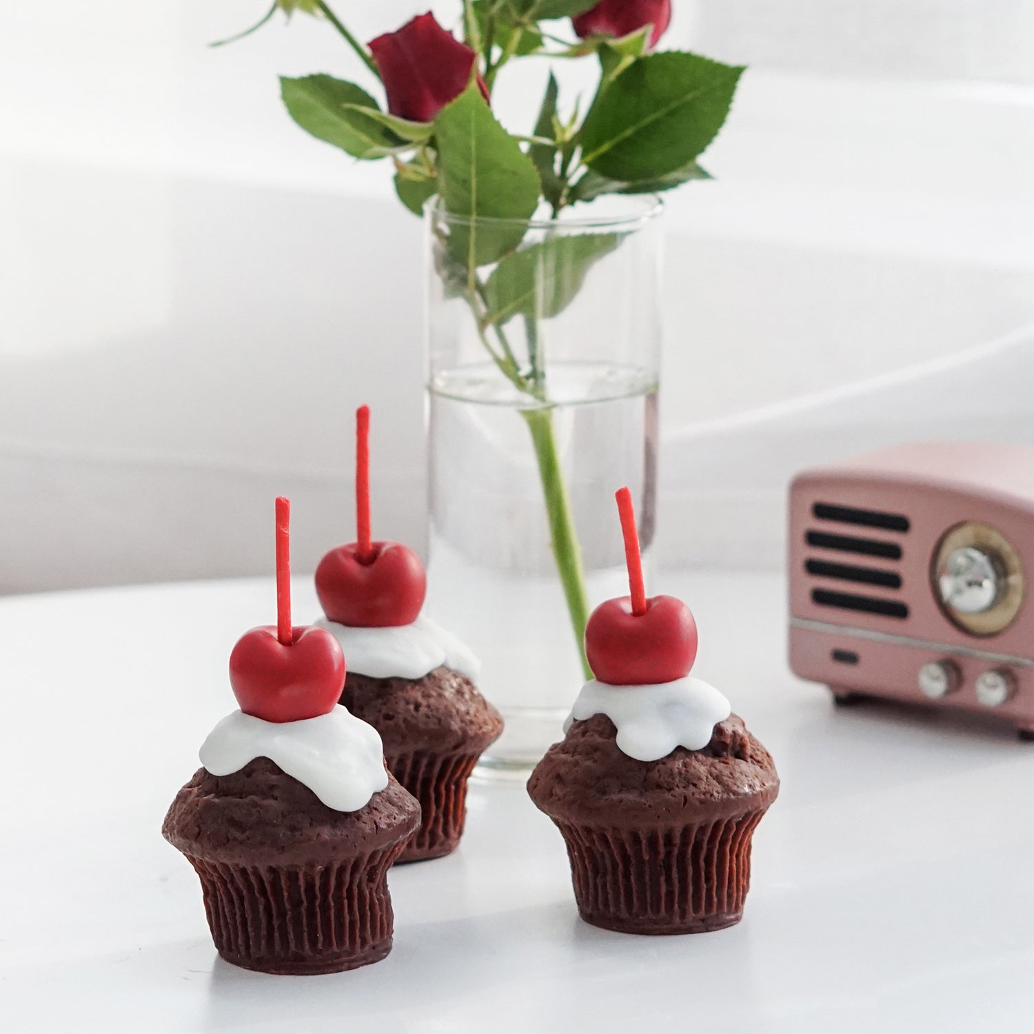 three chocolate cherry muffin shape soy pillar candles, red roses in a clear cylinder flower vase, and pink mini speaker on white table