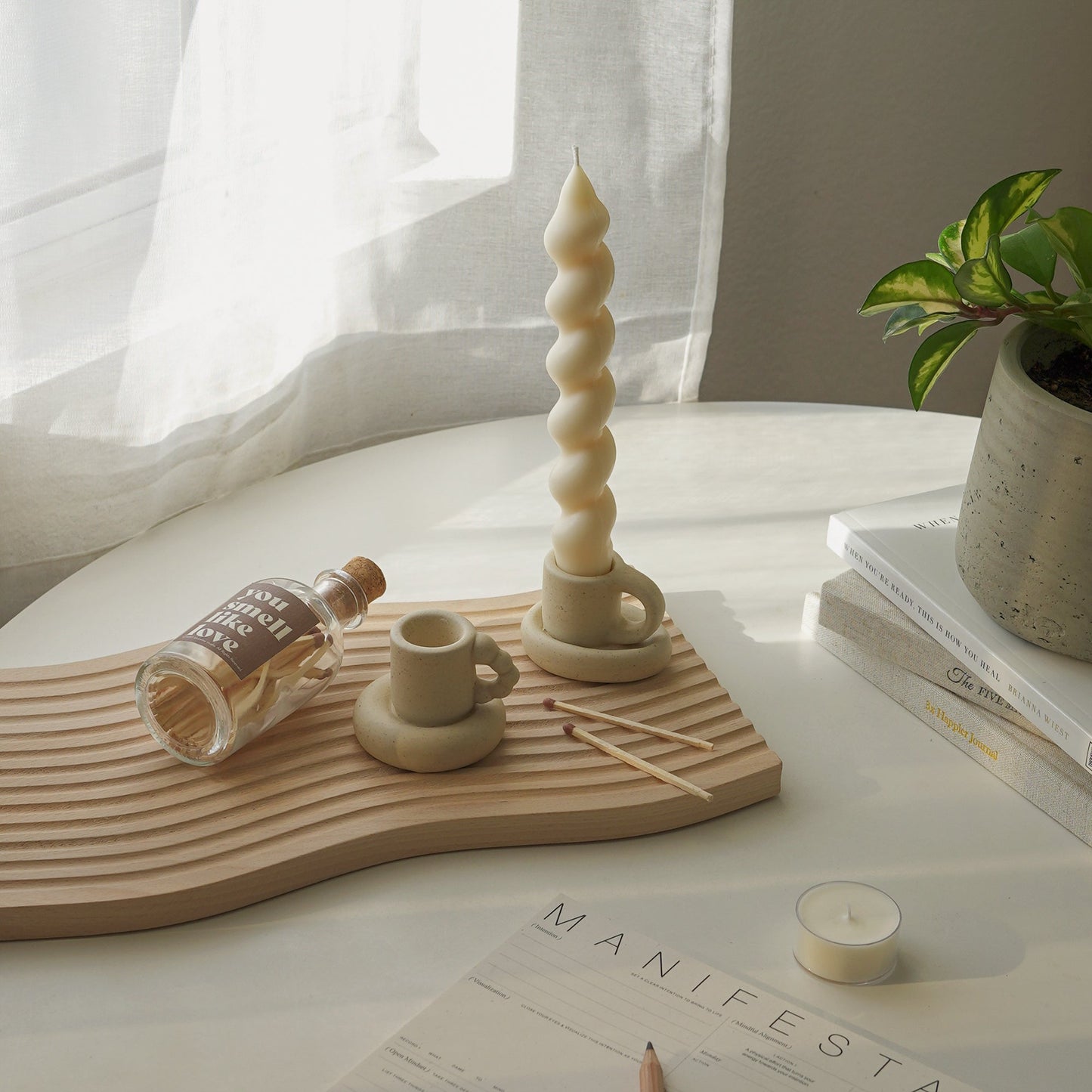 bubble mug candle holder, a wavy candle in a chunky mug candle holder, matches, and a match bottle with a brown sticker inscribed with you smell like love on carved wave wooden tray, a tealight candle, manifestation note, and plant pot placed on books on the white round table