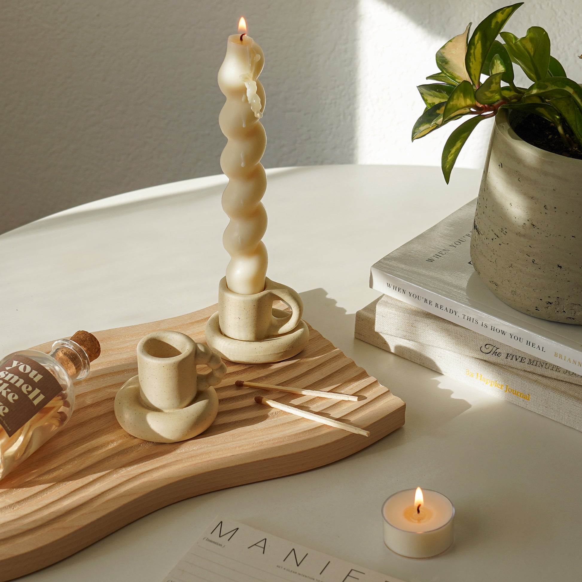 bubble mug candle holder, a wavy candle in a chunky mug candle holder, matches, and a match bottle with a brown sticker inscribed with you smell like love on carved wave wooden tray, a lit tealight candle, manifestation note, and plant pot placed on books on the white round table