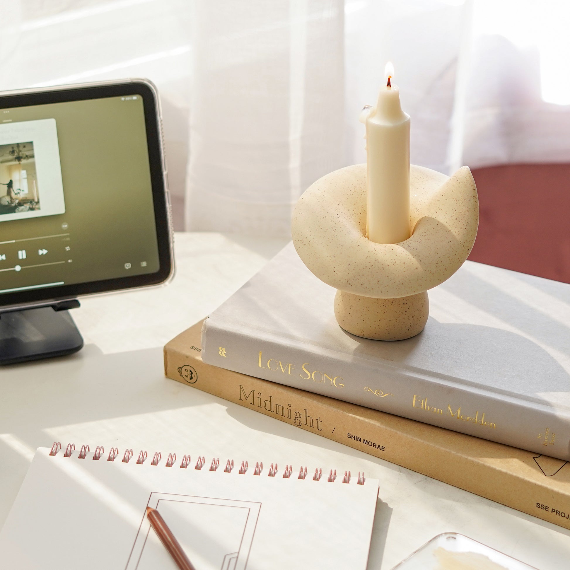 a lit crayon shape white soy pillar taper candle in a dotted beige ceramic mushroom candle holder placed on books love song and coloring book called midnight by shin morae, sketchbook, color pencil, and ipad mini playing music london by yerin baek