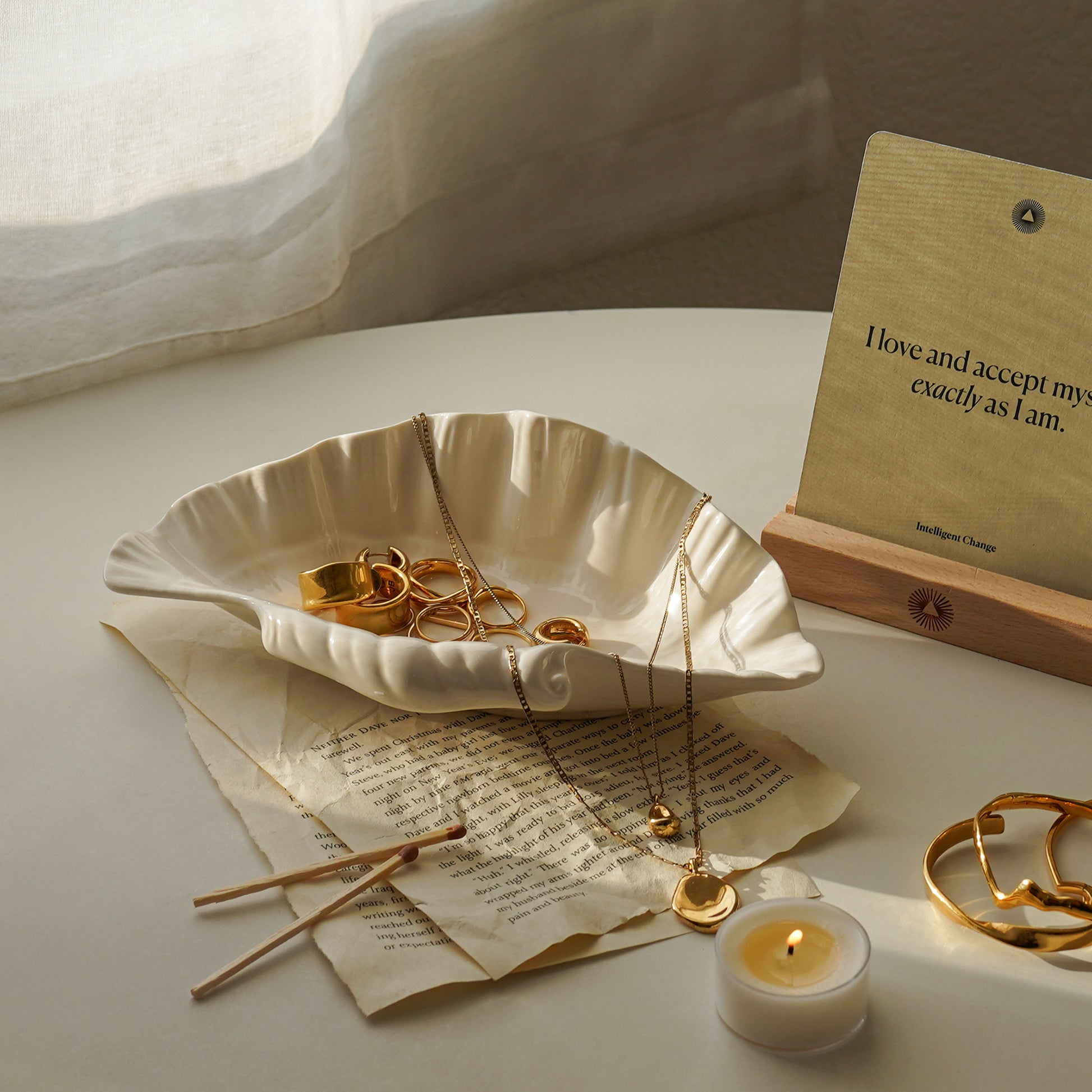 gold jewelry placed in an open shell ceramic tray on book pages, matches, a lit tealight candle, gold bracelets, and affirmation card on the white round table