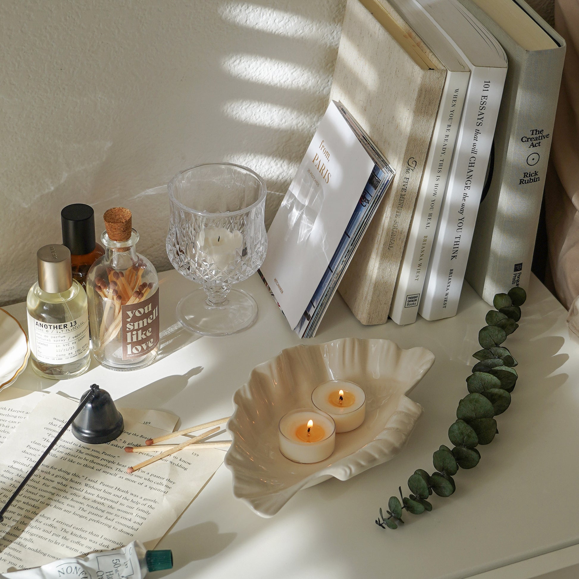 two tealight candles burning in a ceramic shell shape tray, books, postcards, match bottle, perfume, and candle snuffer on white shelves by the bed