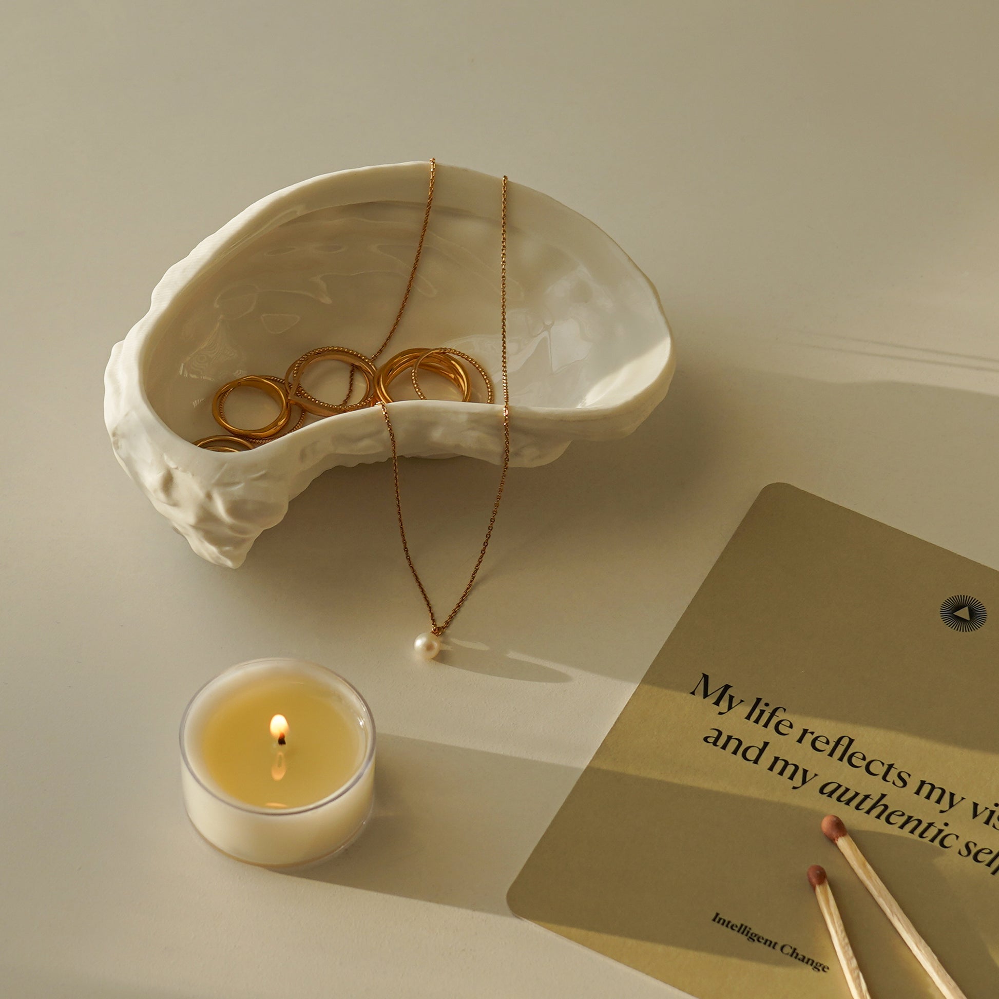 gold jewelry in an oyster dish, a lit tealight candle, and affirmation card from Intelligent Change on the white table