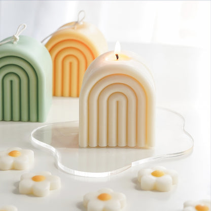 lit white rainbow soy pillar candle on a clear wavy acrylic coaster, sage green and tangerine yellow rainbow shaped sculptural soy pillar candles with long cotton wicks, and daisy flower shape wax melts on the white table