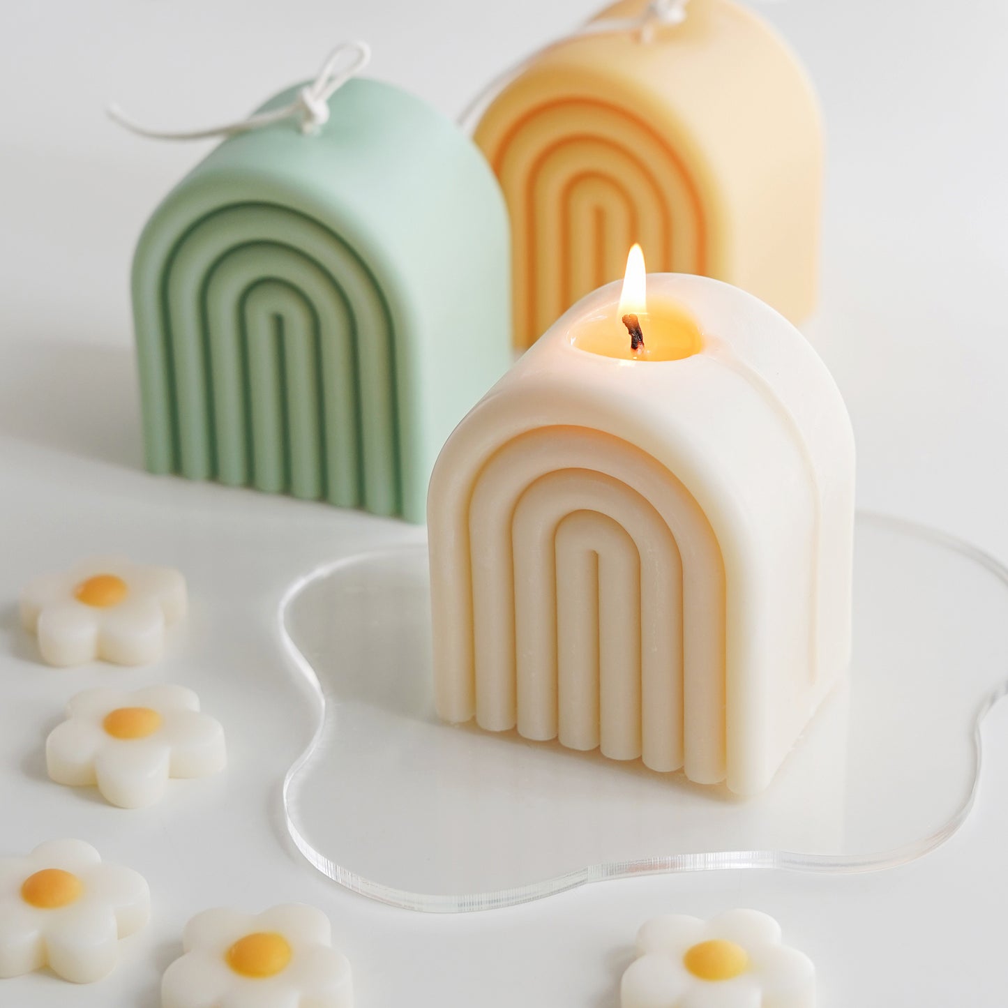 lit white rainbow soy pillar candle on a clear wavy acrylic coaster, sage green and tangerine yellow rainbow shaped sculptural soy pillar candles with long cotton wicks, and daisy flower shape wax melts on the white table