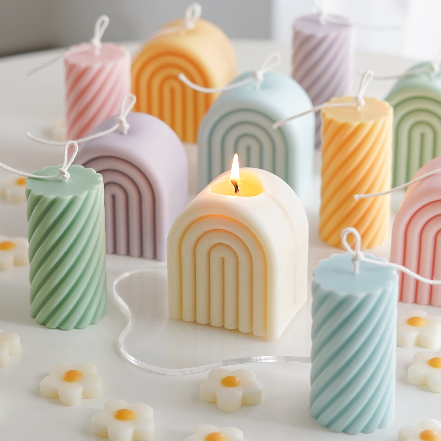 lit white rainbow soy pillar candle on a clear wavy acrylic coaster, sage green, lavender, aqua blue, tangerine yellow, blush pink rainbow shaped soy pillar candles and spiral sculptural candles with long cotton wicks, and daisy flower shape wax melts on the white table