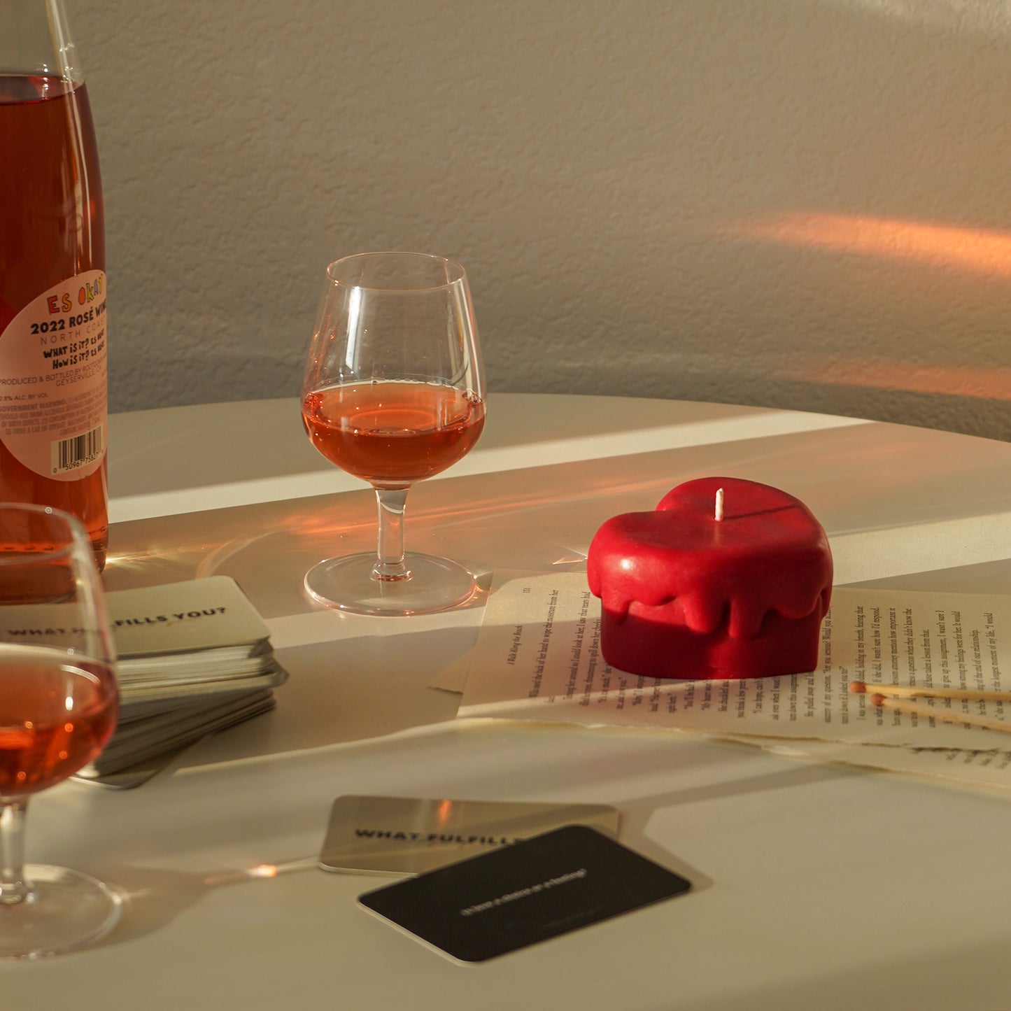red melting heart candle and matches on book pages, a glass of rose wine, a bottle of rose and card on the white round table