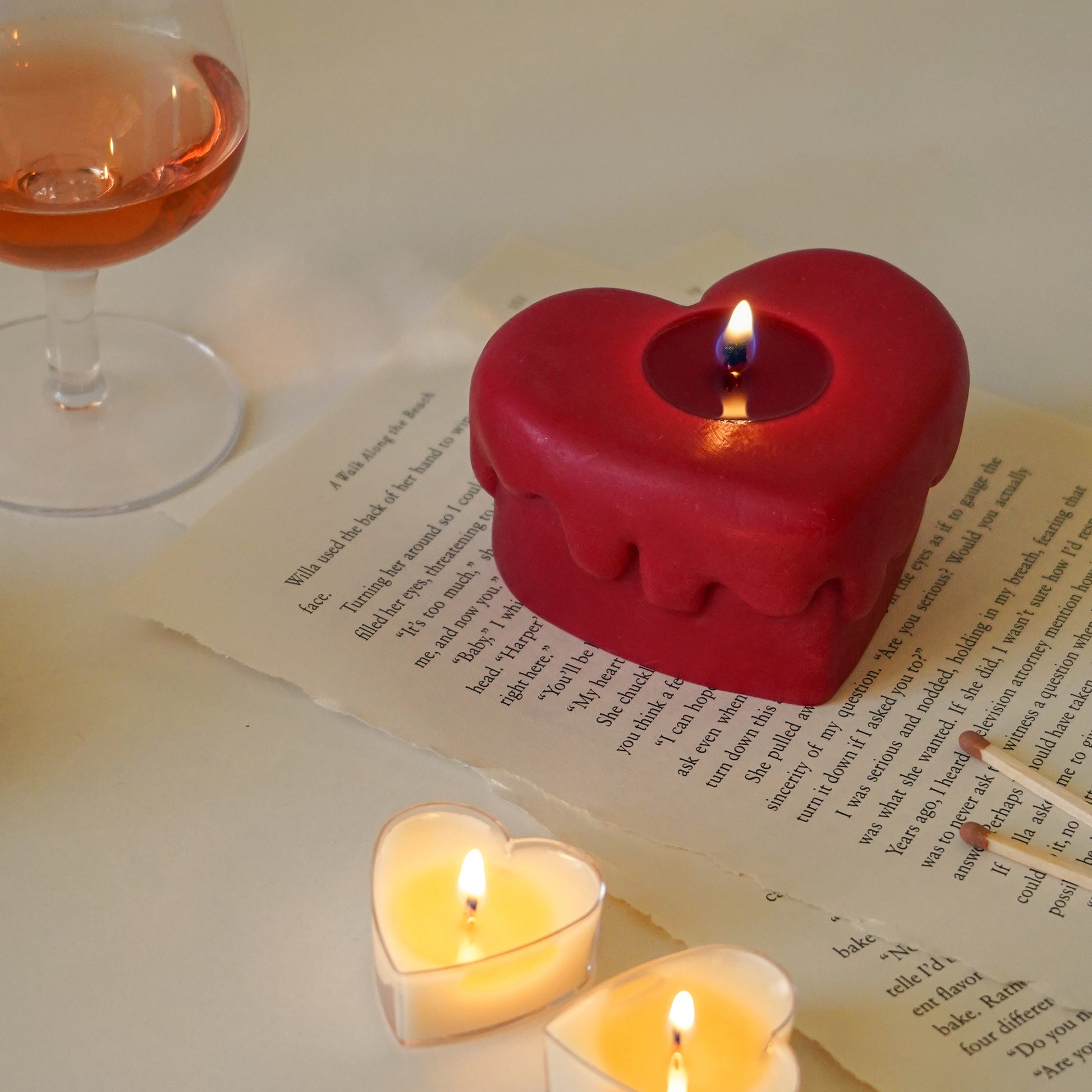 red melting heart candle on book pages, a glass of rose wine and two heart tealight candles