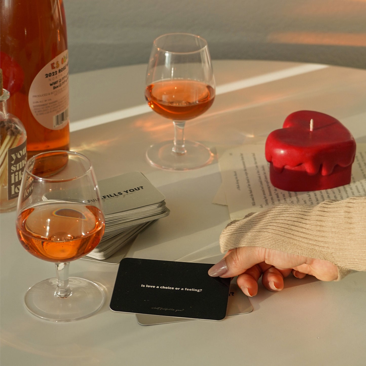 hand holding cards and red melting heart candle and matches on book pages, a glass of rose wine, and a bottle of rose on the white round table
