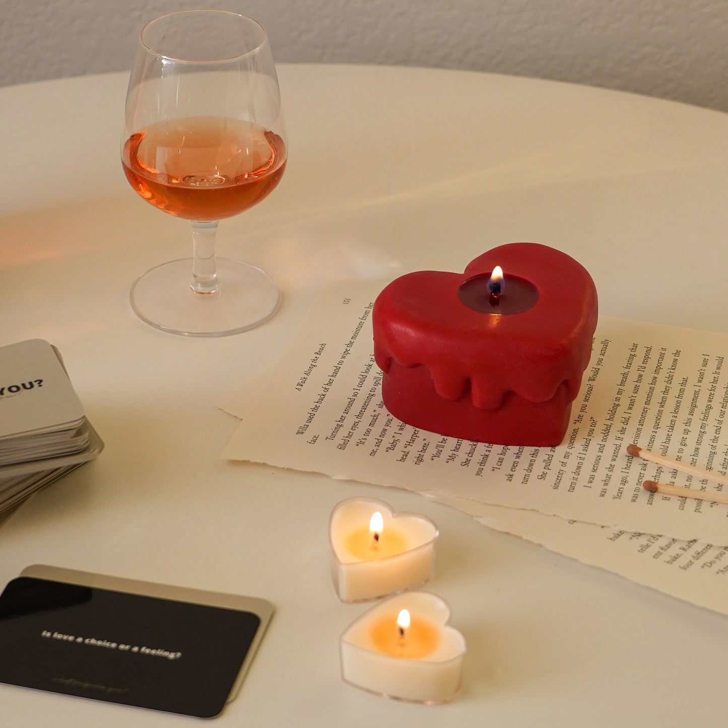 red melting heart candle on book pages, a glass of rose wine and two heart tealight candles
