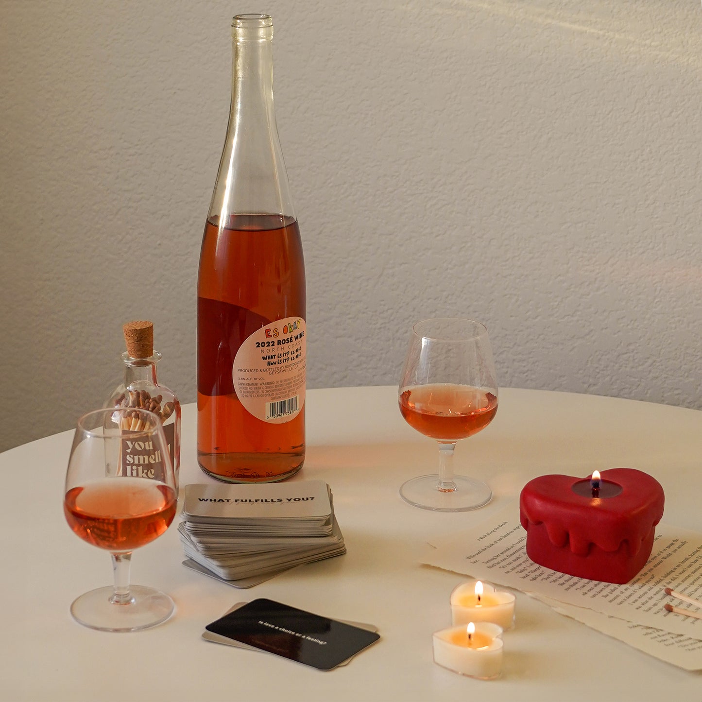 red melting heart candle and matches on book pages, a glass of rose wine, a bottle of rose and card on the white round table