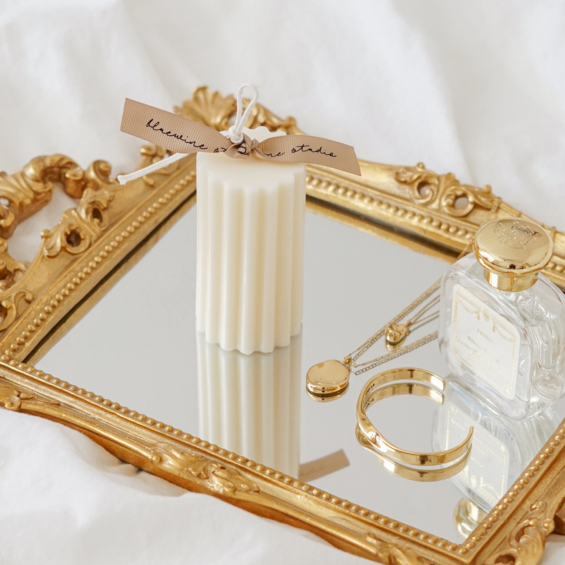 a white fluted ribbed cylinder soy pillar candle with beige bluewine stuio, jenny bird necklace and bracelet, and santa maria novella on a rectangular french gold mirror tray placed on a sheet of bed