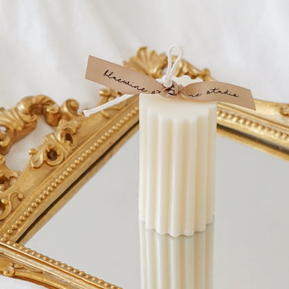 a white fluted ribbed cylinder soy pillar candle with beige bluewine stuio placed on a rectangular french gold mirror tray placed on a sheet of bed