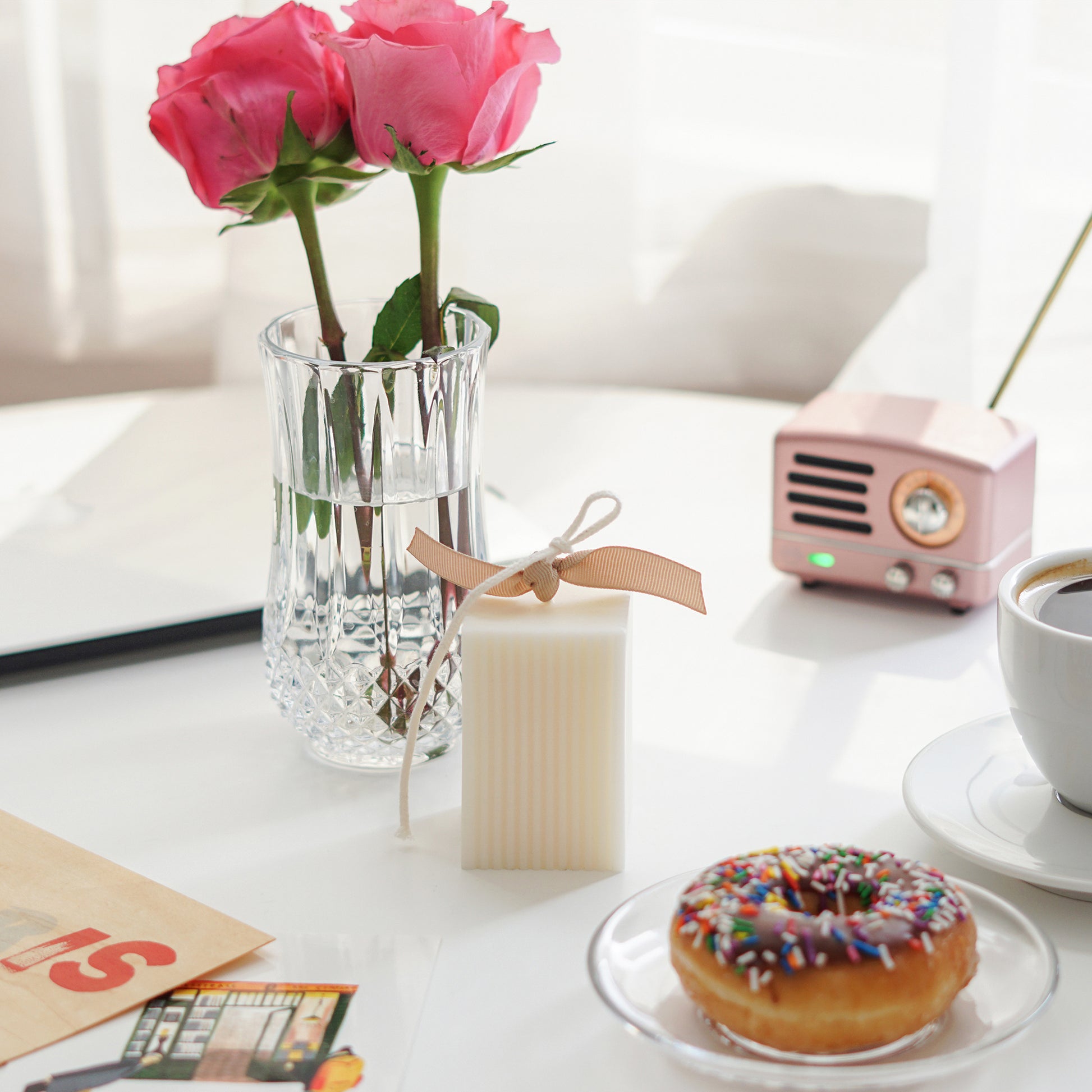 pink roses in a glass, ribbed rectangular white soy pillar candle, chocolate sprinkle donut on a clear plate, cup of coffee in a illy mug, pink mini speaker, paris wooden postcard, macbook on white round table