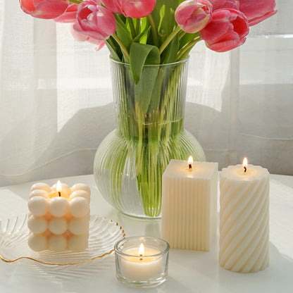 a lit square cube bubble soy pillar candle on a gold rim clear shell tray, a lit tealight candle in a glass candle holder, a lit ribbed square pillar soy candle, a lit spiral white soy pillar candle, and pink tulips in a fluted vase