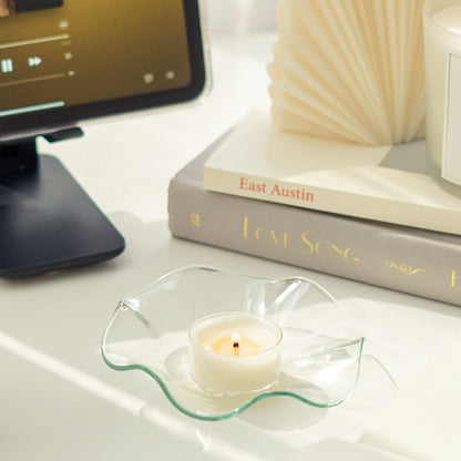 a lit tealight candle on a clear wavy ruffle dish, starbucks tumbler, pencil , and a lit frosted glass container soy candle on books