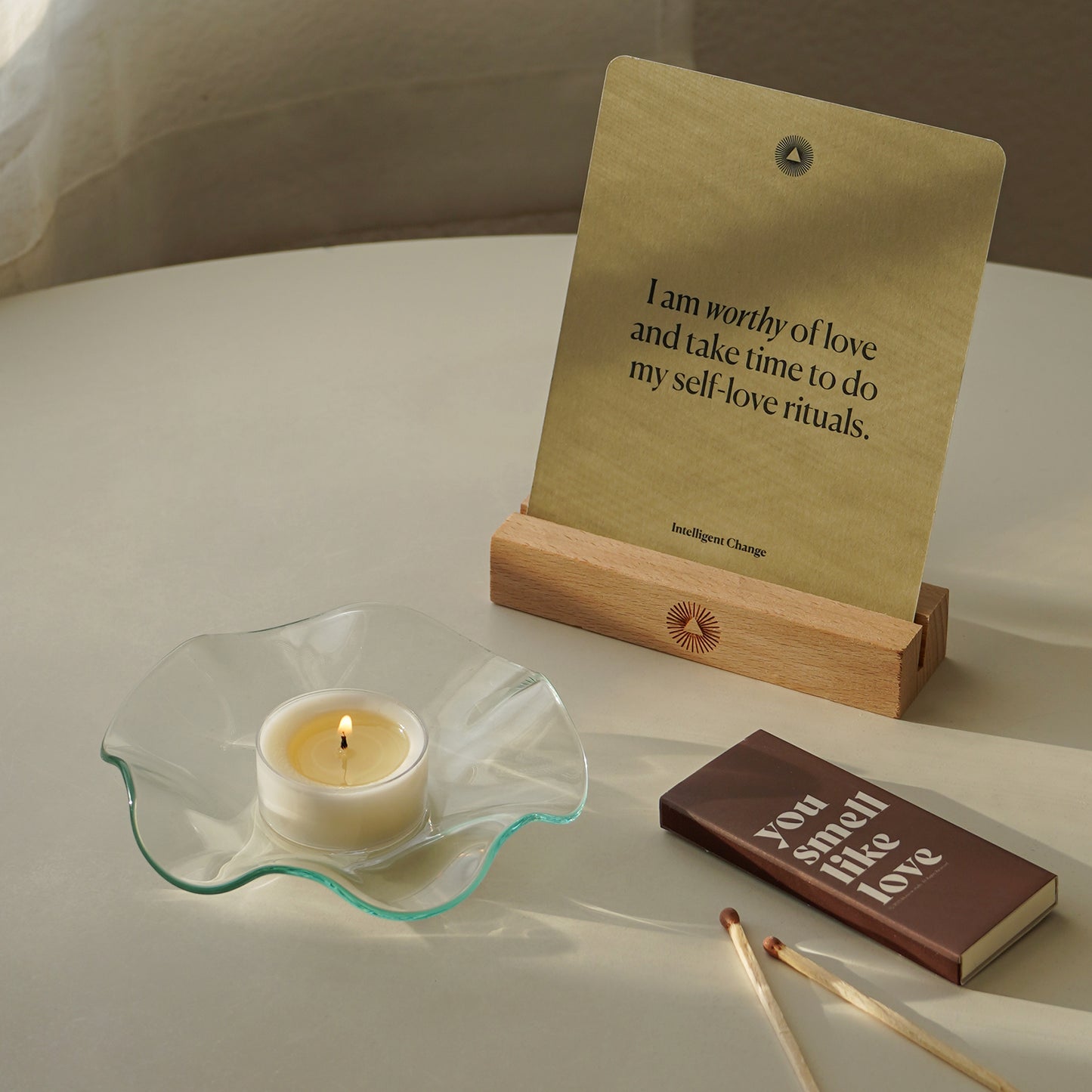 a lit tealight candle placed on a clear ruffle dish, brown matches, a brown matchbox inscribed with "you smell like love" and an affirmation card on the white round table