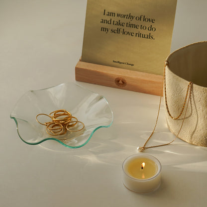 gold rings on clear ruffle dish, a lit tealight candle, ceramic mug with gold necklace, and an affirmation card