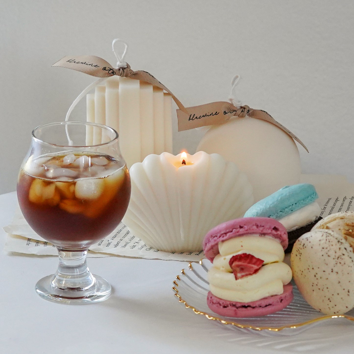 three macarons on a gold rim seashell clear tray, iced americano in a mini glass, a lit seashell soy pillar candle, ribbed stair candle and round sphere candle with beige bluewine studio ribbons