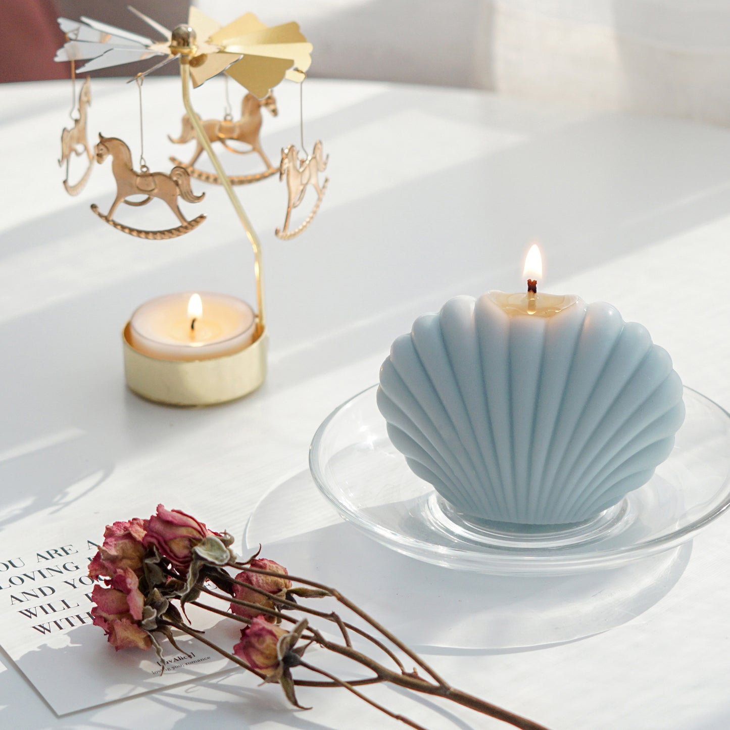 a lit aqua blue seashell shape soy pillar candle on a clear dish, pink dried roses, a postcard, and a lit tealight candle in a gold merry-go-round tealight candle holder on white round table