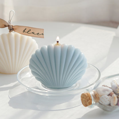 a lit aqua blue seashell shape soy pillar candle on a clear dish, and a white seashell candle with beige bluewine studio ribbon
