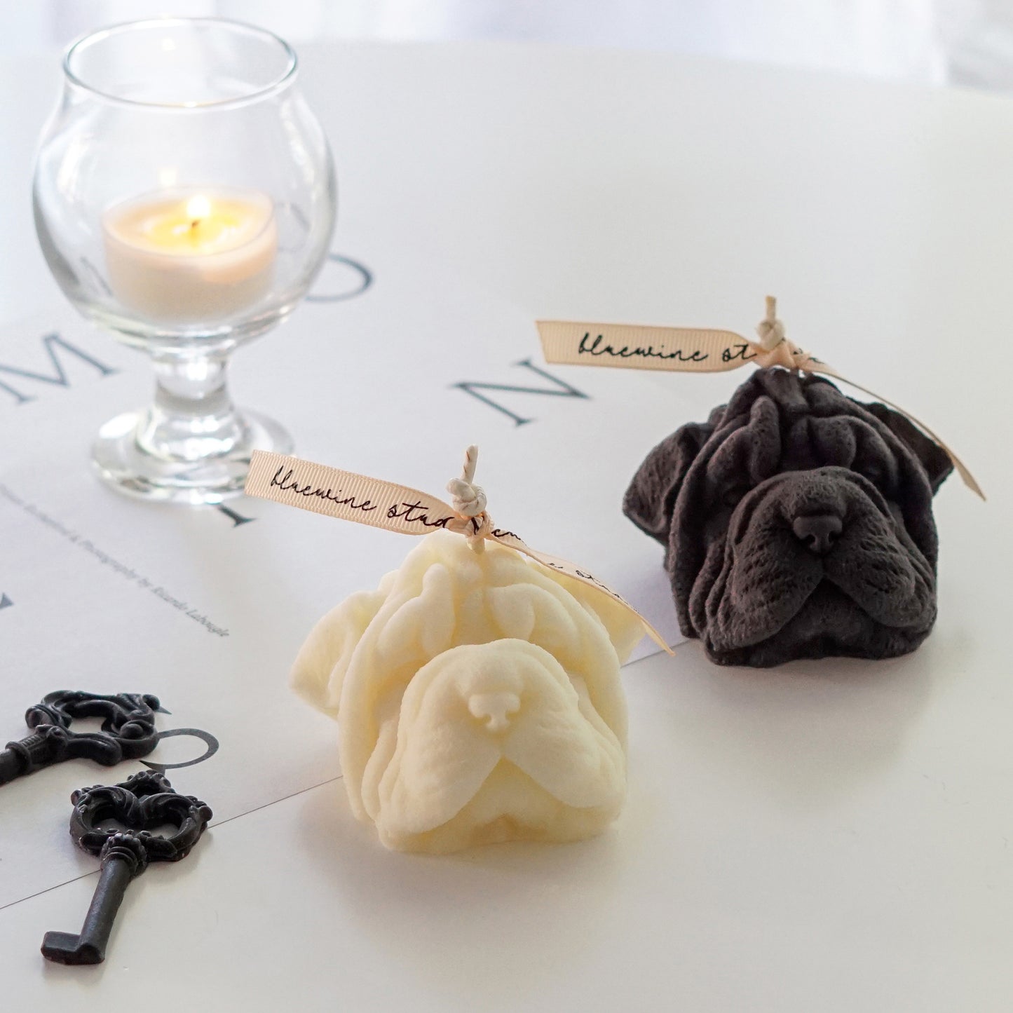 cream and dark gray shar pei shape dog soy pillar candles with bluewine studio ribbon, black key shape wax melts, a lit tealight candle in a mini glass