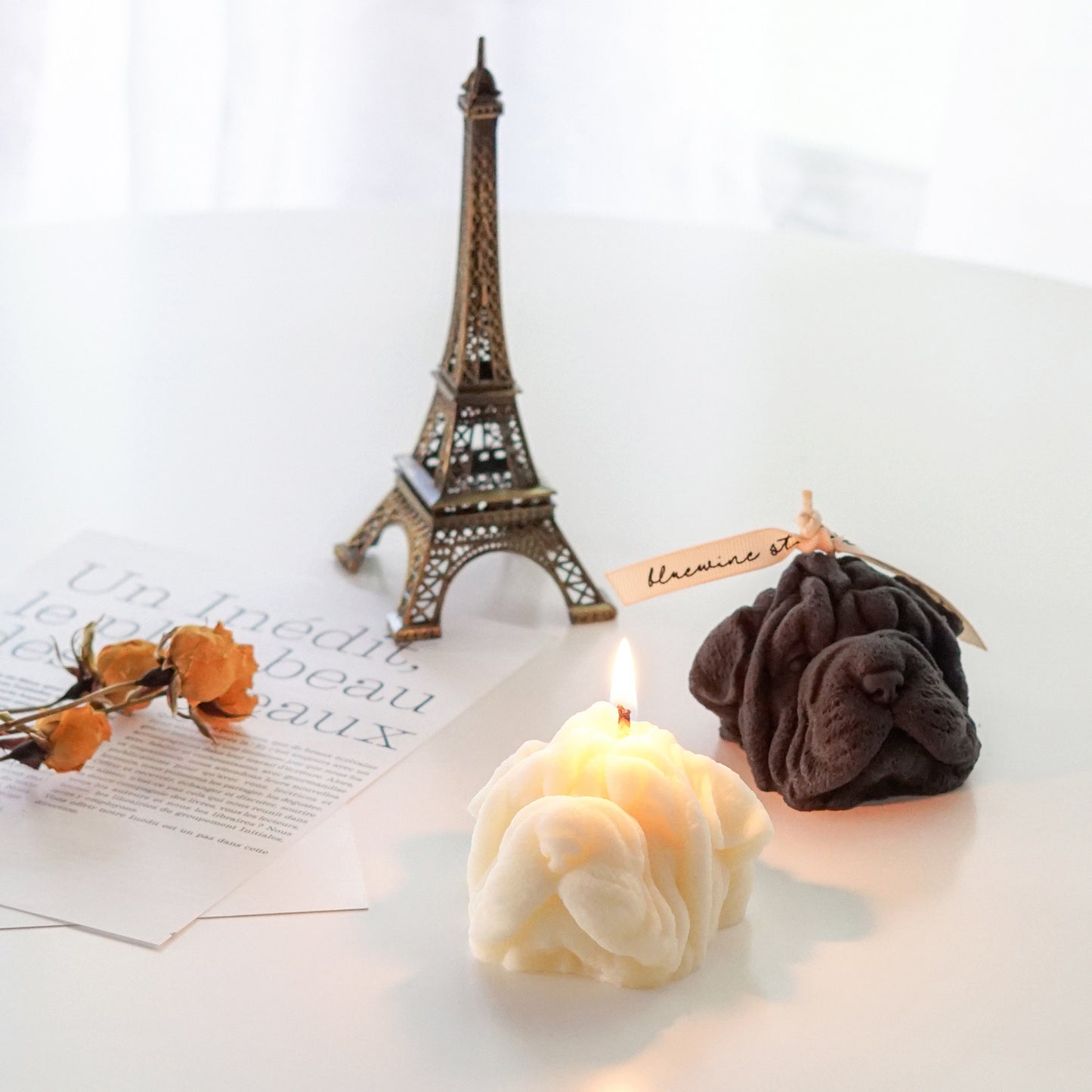 a lit cream shar pei soy candle, dark gray shar pei candle, yellow dried roses, magazine papers, and mini eiffel tower home decor
