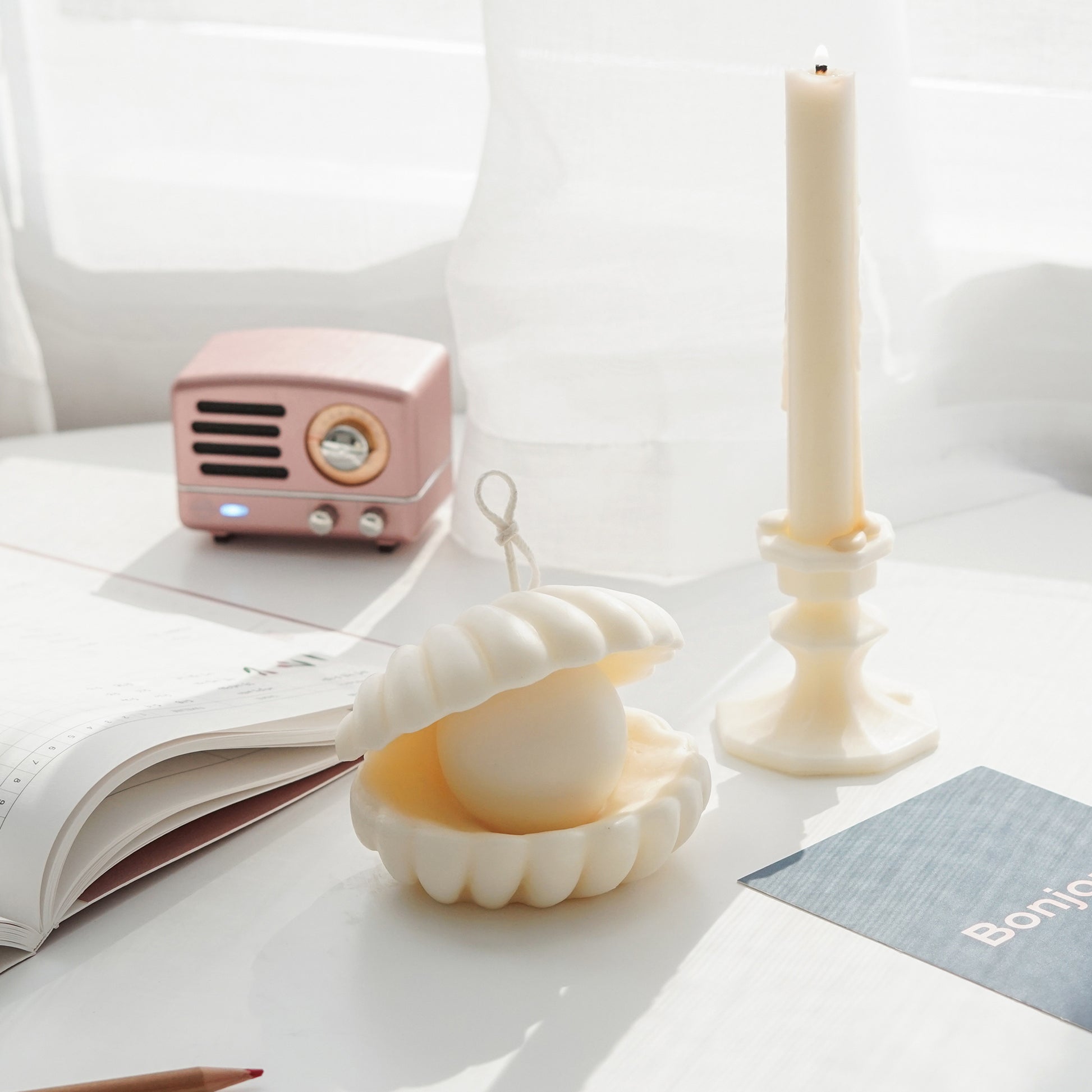 shell pearl white soy pillar candle, blue bonjour postcard, planner, color pencil, pink mini speaker, and a lit taper candle